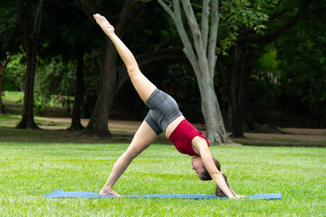 Stunning hispanic woman wearing workout clothing stretching and practicing yoga in a public park. Three legged dog pose or Eka Pada Adho Mukha Svanasana.