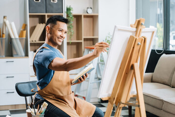 Asian male painter do artwork in art workshop, painting supplies, oil pastels, two canvas easel, creative space with paintbrush in art studio