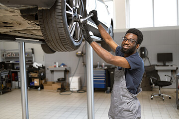 Smiling mechanic in overalls working on car in garage. Automotive technician using tools for car maintenance and repair. Modern workshop with equipment and tools.