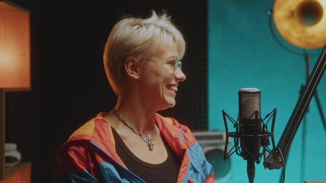 Young joyous woman with short blonde hair wearing colorblock jacket smiling and talking into microphone as recording podcast interview in audio studio