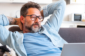 One adult man outstretching arms looking an open laptop sitting comfy on the couch at home. End work or browsing internet lazy afternoon concep tlifestyle people. Modern worker indoor apartment.