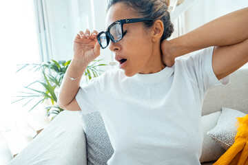 Close up front view of adult woman yaws at home tired and exhausted siting on sofa. Portrait of female ready to sleep or just woke up from afternoon nap. Energy needed lady wearing glasses alone