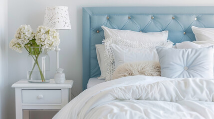 Cozy bedroom decorated in whites and blues. White bed linens and fluffy pillows rest on a blue headboard. A bedside table completes the serene atmosphere.