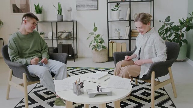 Full arc shot of female Caucasian sign language tutor and deaf teenage male student with cochlear implant sitting together during private lesson, having conversation with sign language and laughing