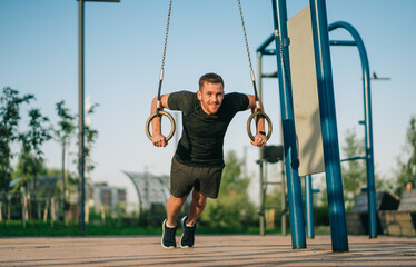 man exercises on gymnastic rings in an outdoor fitness park. He is mid-pose, leaning forward and smiling, with greenery and blue skies in the background