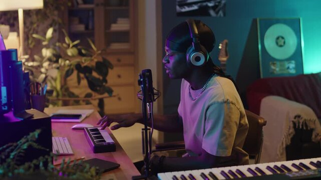 Black musician wearing headphones playing keyboard, using computer and singing in mic in home recording studio illuminated by colorful neon lights