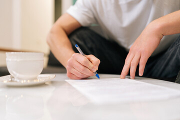 Closeup hands of unrecognizable male client signing papers sitting at table in waiting room. Young man with pen putting signature on medical card official contract agreement, writing documents.