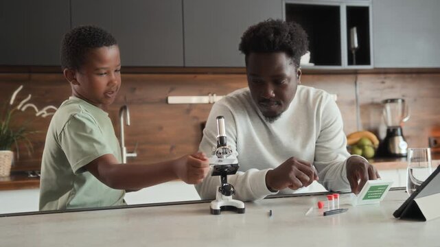 African American boy and man is sitting at table in kitchen, studying and using microscope together. Black skinned son and father spend time researching chemistry at home.