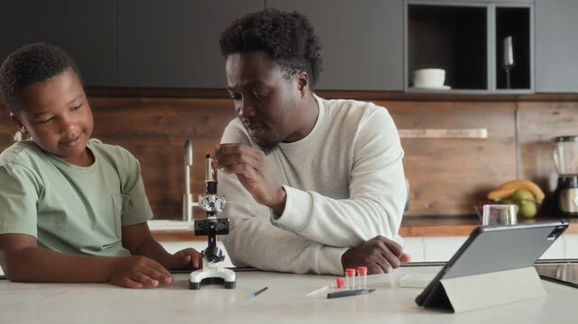 African American boy and man is sitting at table in kitchen, studying and using microscope together. Black skinned son and father spend time researching chemistry at home.