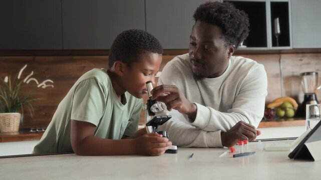 African American boy and man is sitting at table in kitchen, studying and using microscope together. Black skinned son and father spend time researching chemistry at home.