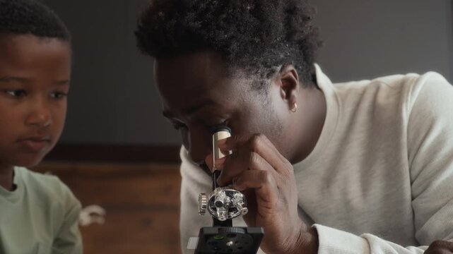 African American boy and man is sitting at table in kitchen, studying and using microscope together. Black skinned son and father spend time researching chemistry at home.