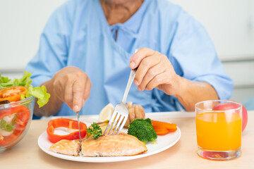 Asian senior woman patient eating Salmon steak breakfast with vegetable healthy food while sitting and hungry on bed in hospital.