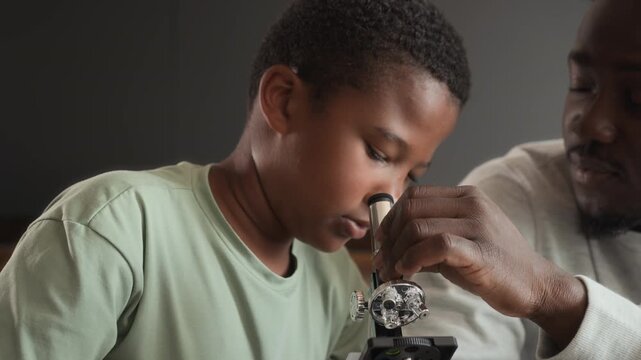 African American boy and man is sitting at table in kitchen, studying and using microscope together. Black skinned son and father spend time researching chemistry at home.