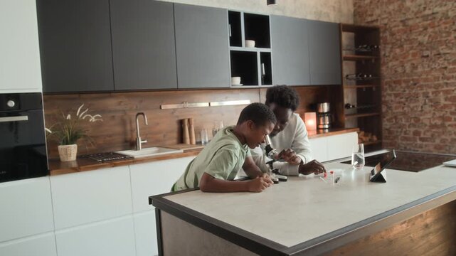 African American boy and man is sitting at table in kitchen, studying and using microscope together. Black skinned son and father spend time researching chemistry at home.