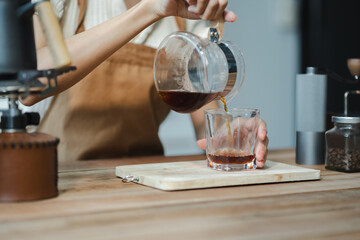 Drip coffee, woman making drip coffee by pouring spills hot water on coffee ground with prepare filter from copper pot to glass transparent chrome drip maker on wooden table.