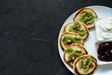 Plate of pancakes dripping with cherry jam, sour cream and kiwi pieces with dark gray background.. Copy space for text. Mockup for menu, Pancake day