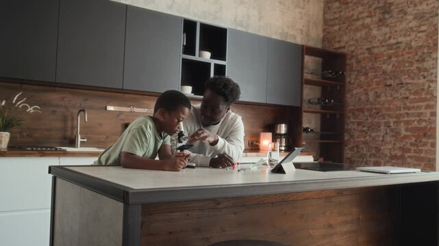 African American boy and man is sitting at table in kitchen, studying and using microscope together. Black skinned son and father spend time researching chemistry at home.