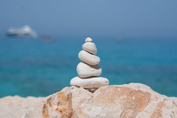 A tranquil stone cairn stands on a rocky shore by the sea, with blurred boats in the background