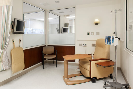 Interior view of a hospital treatment cubicle with a chair and a drip treatment stand, trolley table and a television.
