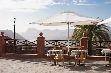 cafe restaurant outdoor terrace with white umbrella table and chairs, with view on palm tree