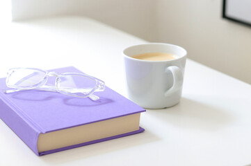 A purple mock up book with transparent glasses and a white cup of coffee on white table. Free copy space for text