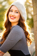 Portrait of a smiling beautiful young woman wearing a grey beret, with her long brown hair back lit by the late afternoon sunlight. 