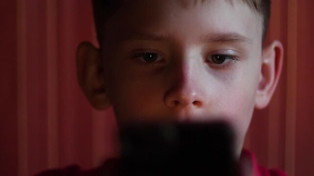 Caucasian boy 9-10 years old looks with horror and bewilderment at the phone screen in the dark, covering his mouth with his hand, obscene content