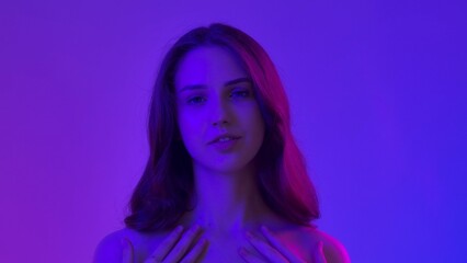 Young woman posing at camera touching smooth skin and smiling, isolated on blue neon background. Natural beauty and skin care concept