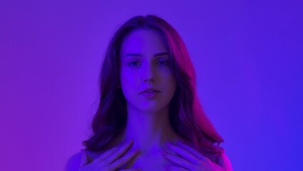 Young woman posing at camera touching smooth skin and smiling, isolated on blue neon background. Natural beauty and skin care concept