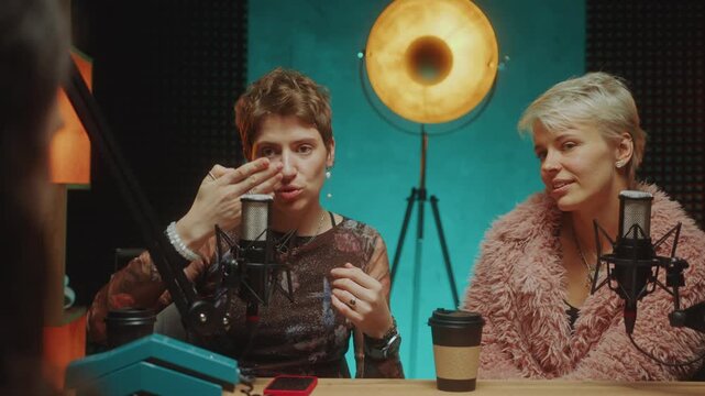 Two young female fashion influencers in trendy outfits having discussion and speaking into microphones when giving interview to podcast host in recording studio