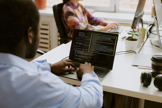 Over shoulder shot of African American man sitting at white wooden table and writing codes while his coworker typing on keyboard in blurred background