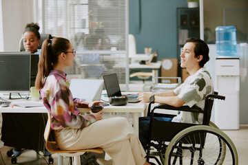 Two Caucasian coworkers having nice conversation while female colleague drinking coffee and her male colleague with disability coding