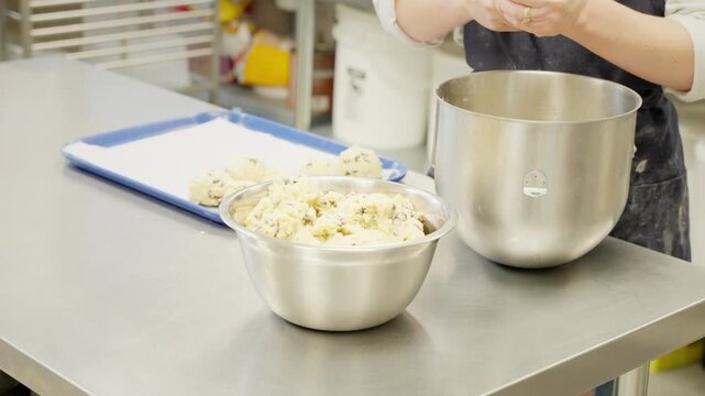 Woman Scoops Cookie Dough from bowl on Stainless Steel Table in Professional Kitchen