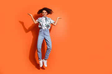 Full length photo of american woman in jeans and summer stylish shirt jumping air comparing two products isolated on orange color background