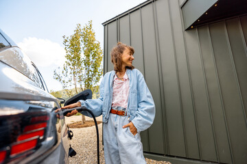 Young woman plugging a charger into electric vehicle near her house outdoors. Concept of modern...