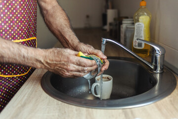 Side view of elderly man with white hair and beard wearing apron washing dishes. Corner of kitchen. Grandfather at work. Bright light from window.