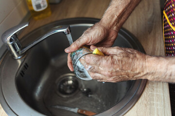 Senior white man standing next to a kitchen sink and rinsing a glass with running water inside a kitchen.