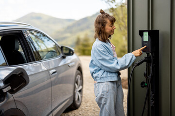 Woman switches on car charging station while charging her electric vehicle near home. Concept of...