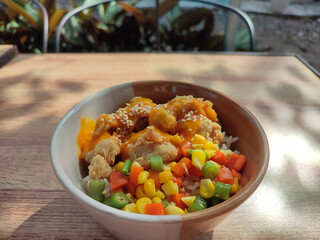 Rice Korean chicken cheese in a bowl on a wooden table with a blurred garden background.    Rice bowl filled with chicken and vegetables.  Negative space.