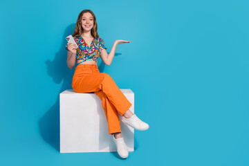 Full size photo of nice young girl sit podium hold phone empty space wear top isolated on blue color background