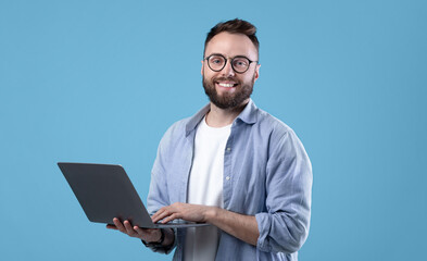 Cheerful millennial guy in glasses using laptop computer for online work or communication on blue studio background. Handsome young man watching video, participating in webinar