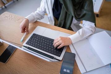 Close-up of a woman in a green hijab working on her laptop with a notebook beside her in a modern office This image emphasizes organization and productivity