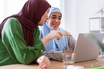 Two women in hijabs discuss a project in a bright office environment They are using a laptop and a notepad highlighting their collaborative efforts and teamwork