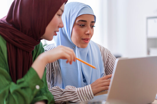 Two women in hijabs collaborate on a project in a modern office space One woman in a green hijab is smiling while the other in a blue hijab focuses on their work