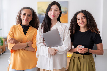 Three confident women stand in a bright office space They are dressed in professional attire and pose confidently for the camera highlighting their professionalism and teamwork