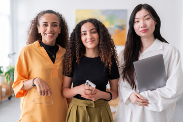 Three confident women stand in a bright office space They are dressed in professional attire and pose confidently for the camera highlighting their professionalism and teamwork