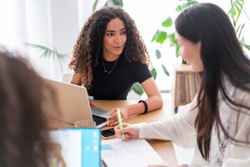 Three young professionals engaged in a team discussion, sharing ideas and working on laptops in a modern, bright office environment with plants in the background