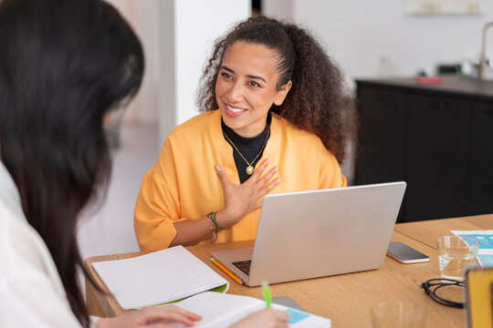 Two professionals having a discussion in a modern office, with one smiling and expressing thoughts while the other listens attentively, with documents and laptops on the table