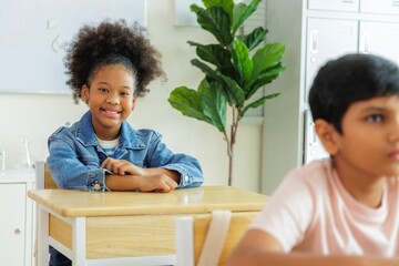 Fototapeta premium Smiling young girl in denim jacket sits at a desk in a classroom. Her classmate is sitting next to her.