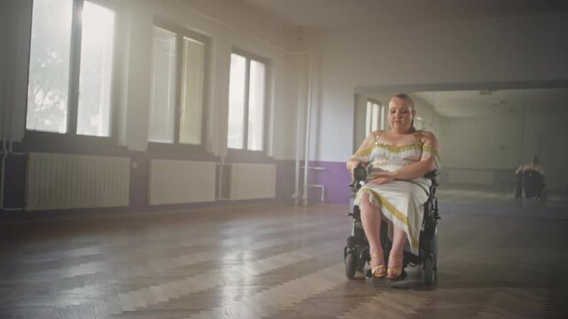 Confident pretty young woman with a disability, driving her electric wheelchair in a dance hall, looking and smiling at a camera, inspiration portrait.